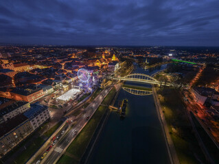 Fototapeta premium A vibrant aerial view of a city at night with illuminated buildings, a glowing bridge, and a ferris wheel by the river. The blue hour sky and reflections on the water create an atmospheric urban scene