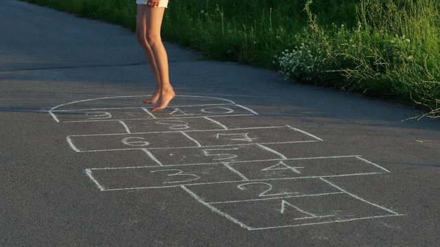 A child jumps barefoot on a hopscotch grid drawn in chalk on the pavement after school. Grass grows nearby in a neighborhood setting