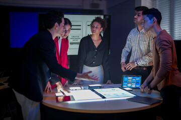 Creative UI/UX design team analyzing mobile app wireframes on illuminated digital touch table. Diverse developers brainstorming web layout strategy in modern high-tech office at night.