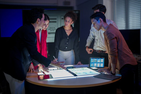 Creative UI/UX design team analyzing mobile app wireframes on illuminated digital touch table. Diverse developers brainstorming web layout strategy in modern high-tech office at night. - Powered by Adobe