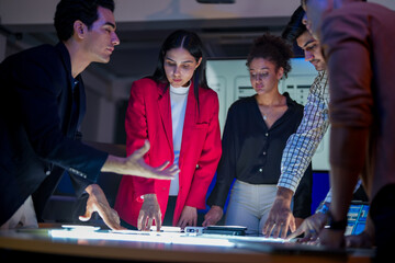 Creative UI/UX design team analyzing mobile app wireframes on illuminated digital touch table. Diverse developers brainstorming web layout strategy in modern high-tech office at night.