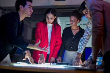 Creative UI/UX design team analyzing mobile app wireframes on illuminated digital touch table. Diverse developers brainstorming web layout strategy in modern high-tech office at night.