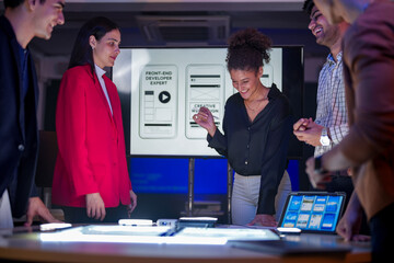 UI/UX design team conducting a workshop to review mobile app prototypes. Front-end developers and visual artists discussing web layout strategy on a large screen in a modern tech startup office.