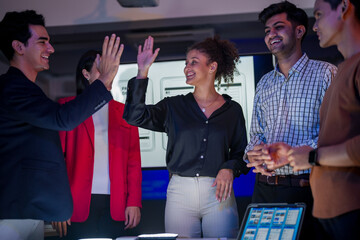 Successful creative team celebrating project achievement in modern office. Happy diverse developers high-fiving after mobile app launch during late night meeting.
