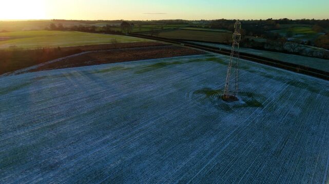 Aerial view of cold frosty fields at sunset, powerlines with golden hour light streaming through, electrical infrastructure and nature, rolling hills in rural England. 