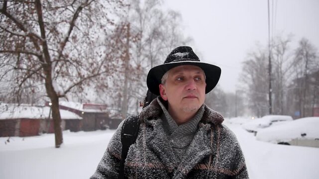 Man in warm clothes with scarf walks along sidewalk among trees and shields himself from heavy snowfall with his hand. Positive mood with smile during extreme winter weather in city.