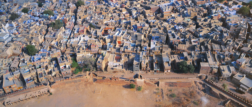 Aerial view of a dense, warm-toned cityscape with blue accents nestled against ancient walls, Jodhpur, Rajasthan, India.