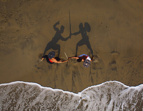 Aerial view of two people casting shadows of warriors engaged in combat on the wet sand near the wavy shoreline, Kozhikode, Kerala, India.