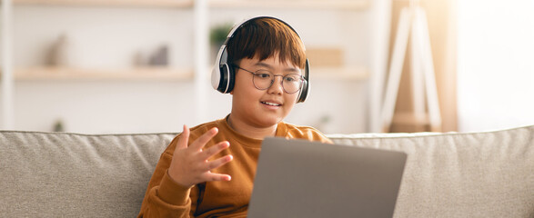 A boy sits on a couch at home, wearing headphones and talking to a laptop. He is engaged in online learning during the afternoon. Light comes in from a nearby window.
