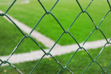 A close-up of a green chain-link fence with a blurred football/soccer field in the background.