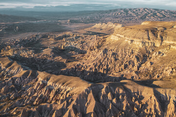 Aerial view of the rugged, sun-kissed landscape of Cappadocia unfolds in a tapestry of warm earthy tones and dramatic rock formations, Cappadocia, Turkey.