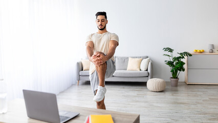 Millennial Arab man stretching his leg in front of laptop during domestic training, following online video tutorial on laptop, copy space. Sporty young guy doing flexibility exercises at home, banner