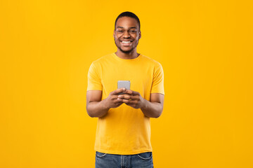 Happy African American Guy Using Phone With New Mobile Application Smiling To Camera Standing Over Yellow Background In Studio. People And Gadgets, Great App For Smartphone Concept