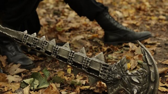 Fallen leaves, Metal halberd, Forest blade. Amidst forest's fallen leaves lies spiked metal weapon, with boots discernible in backdrop.