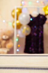Happy birthday concept. Focus on the foreground. A festive blurred background with balloons, a sparkly baby dress on a hanger, and glowing fairy lights. 