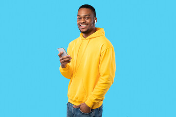Happy African American Guy Using Smartphone Wearing Earbuds Earphones Listening To Music Posing Smiling To Camera Over Blue Studio Background. Male Student Listens To Educational Podcast Online
