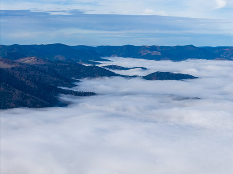 Aerial view of a sea of clouds swallowing the dark, forested peaks, creating a dreamy, ethereal landscape under a pale sky, Thann, Grand Est, France.