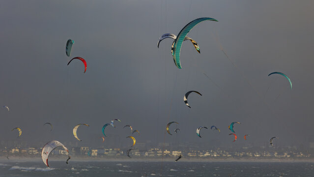 Aerial view of kitesurfers dance on the waves beneath a brooding sky, their colorful kites punctuating the muted tones of the sea and shore, Cape Town, Western Cape, South Africa.