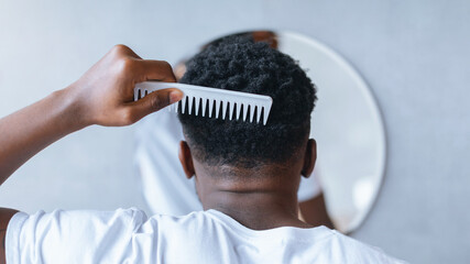 A person stands in front of a round mirror. They are combing their hair with a white comb. The setting is simple with a light-colored wall.