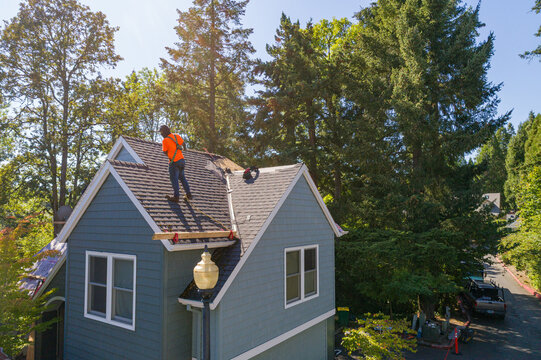 Aerial view of a worker in an orange vest repairing a roof with blue shingles under the sun, Southwest Neptune Court, Beaverton, Oregon, United States.