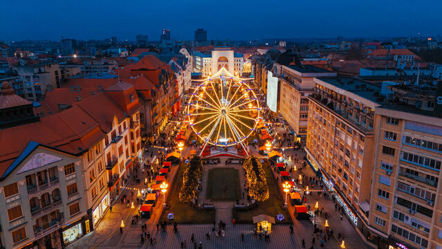 Aerial view of a brightly lit Ferris wheel casting a warm glow over Victory Square and its bustling Christmas market, Timisoara, Timis, Romania.