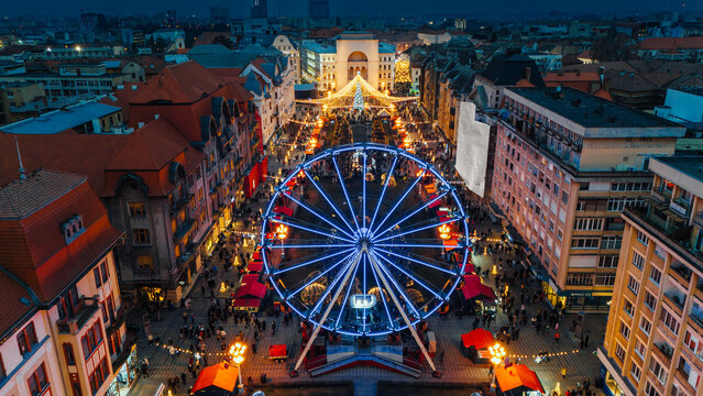 Aerial view of the vibrant Christmas market glowing with festive lights around the Ferris wheel, set against the backdrop of Victory Square, Timisoara, Timis, Romania.