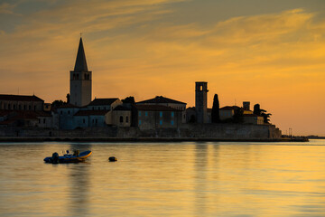 Porec, Croatia - August 13, 2025: Golden sunset over calm water with a historic stone skyline, silhouettes of towers and trees, and a small boat drifting near the peaceful shore.