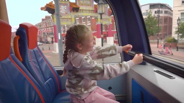 Full of imagination, a young girl playfully pretends to drive from the front seat of a double-decker bus. Through the window, the unique architecture of Liverpool's Chinatown passes by
