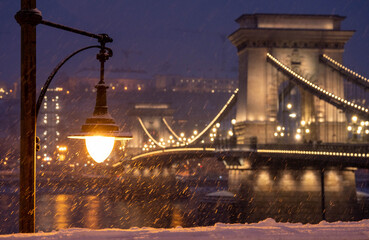Fototapeta premium Budapest, Hungary - January 6, 2026: The historic Szechenyi Chain Bridge glows with golden lights during a heavy evening snowfall in Budapest, framed by a vintage street lamp and snow-covered