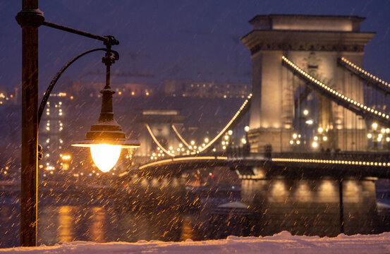 Budapest, Hungary - January 6, 2026: The historic Szechenyi Chain Bridge glows with golden lights during a heavy evening snowfall in Budapest, framed by a vintage street lamp and snow-covered