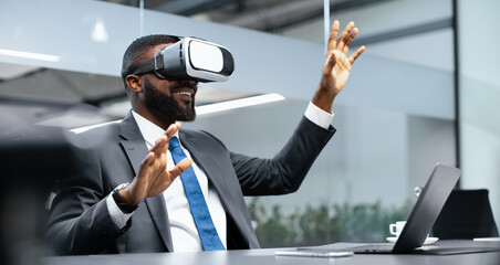A man is wearing a virtual reality headset while sitting at a desk in an office. He appears to be...