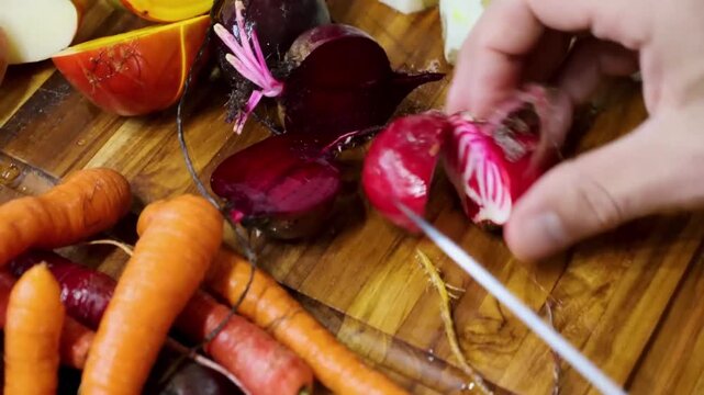 A hand with a knife slices beet and radish into smaller pieces on a wooden board surrounded by other colorful root vegetables.