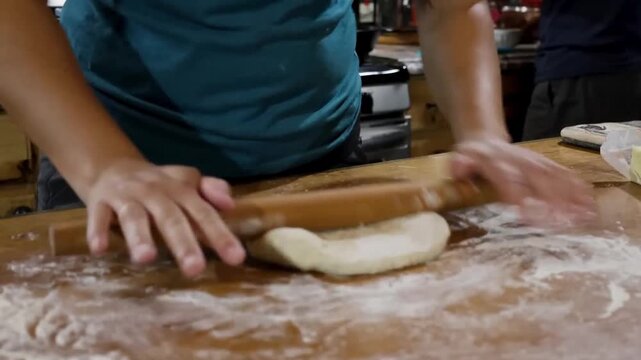 Hands flattening fresh pastry dough with a rolling pin on a wooden kitchen counter.