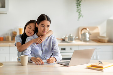 Family business. Young asian woman working on laptop and taking notes, loving cute daughter embracing mother, spending time together at home, sitting in kitchen interior