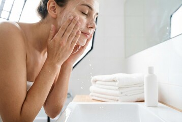 Woman washes her face in bathroom during morning routine while standing at sink with towels in background