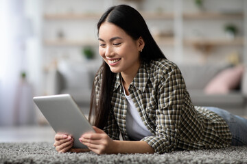 Positive asian woman surfing on social media, using digital tablet, living room interior, copy space. Happy young korean lady laying on floor and reading blog on modern pad, enjoying new mobile app
