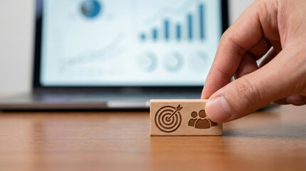 A hand places a wooden block with target and teamwork icons on a desk. A laptop displays graphs and data in the background. This scene represents digital strategy and business growth