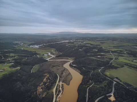 Aerial view of the river snaking through lush green fields and hills under a vast, cloudy sky, Odeceixe, Faro District, Portugal.
