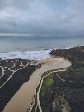 Aerial view of the winding river meeting the sea's foamy embrace amidst the rugged coastline, Praia de Odeceixe, Faro District, Portugal.