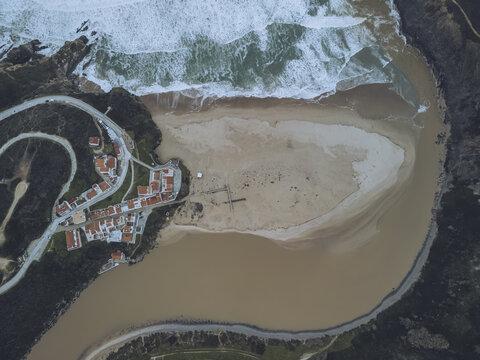 Aerial view of the meeting of the muddy river and the ocean's white waves beside the village with red roofs, Odeceixe, Faro District, Portugal.