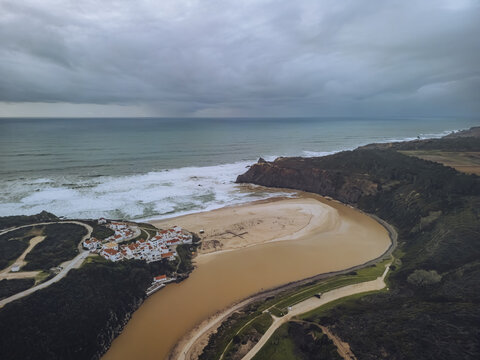 Aerial view of the river snaking towards the ocean, meeting the sandy beach near a cluster of white buildings under a grey, brooding sky, Odeceixe, Faro District, Portugal.