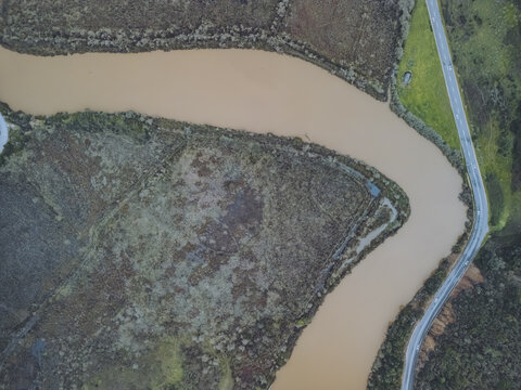 Aerial view of a winding, muddy river cutting through a landscape of dark, textured earth and green vegetation, bordered by a narrow road, Odeceixe, Faro District, Portugal.