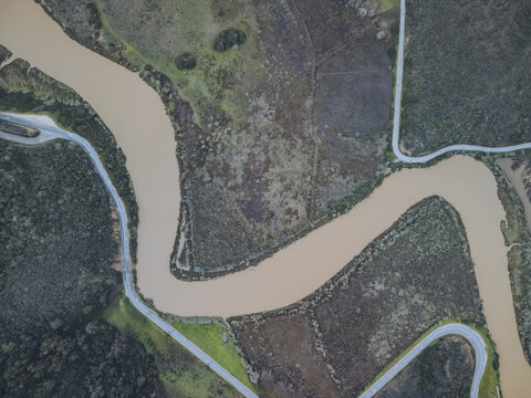 Aerial view of a winding, muddy river snaking through a tapestry of green and brown fields, divided by slender roads, Odeceixe, Faro District, Portugal.