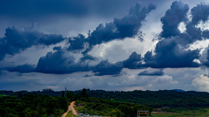 The image shows a landscape of lush green hills and forests under a dramatic, cloud-laden sky, with areas of open land and what appears to be a column of rain in the distance.