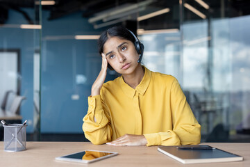 Tired young Indian woman in a suit sitting at a desk in the office and holding her head
