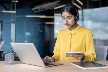 Serious young Indian woman in a suit sitting at a desk, working on a laptop and holding a notebook