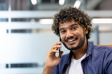 Close-up photo of a young Indian man sitting in an office and smiling while talking on the phone