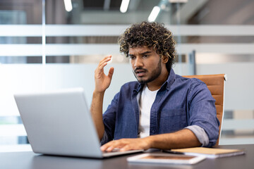 Shocked Indian young man sitting at office desk and looking worriedly at news and problem on laptop