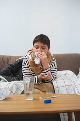 Sick child sitting on sofa wrapped in blanket, holding tissue to nose, winter illness concept, home interior, cold or flu symptoms.