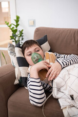 Boy wrapped in blanket using inhaler mask, sitting on sofa at home, recovering from illness, glass of water nearby, cozy indoor setting.
